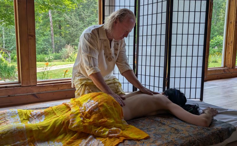 Sergio kneeling in a sunlit room with large windows, his hands on the back of a client who lies face down under a yellow sarong